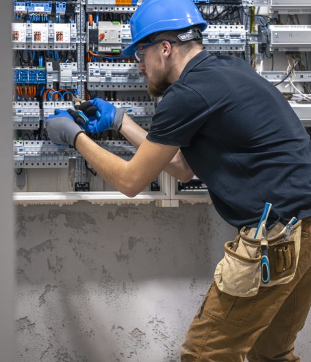 A male electrician works in a switchboard with an electrical connecting cable. Electrician with screwdriver tightens electrically operated switching equipment in fuse box.